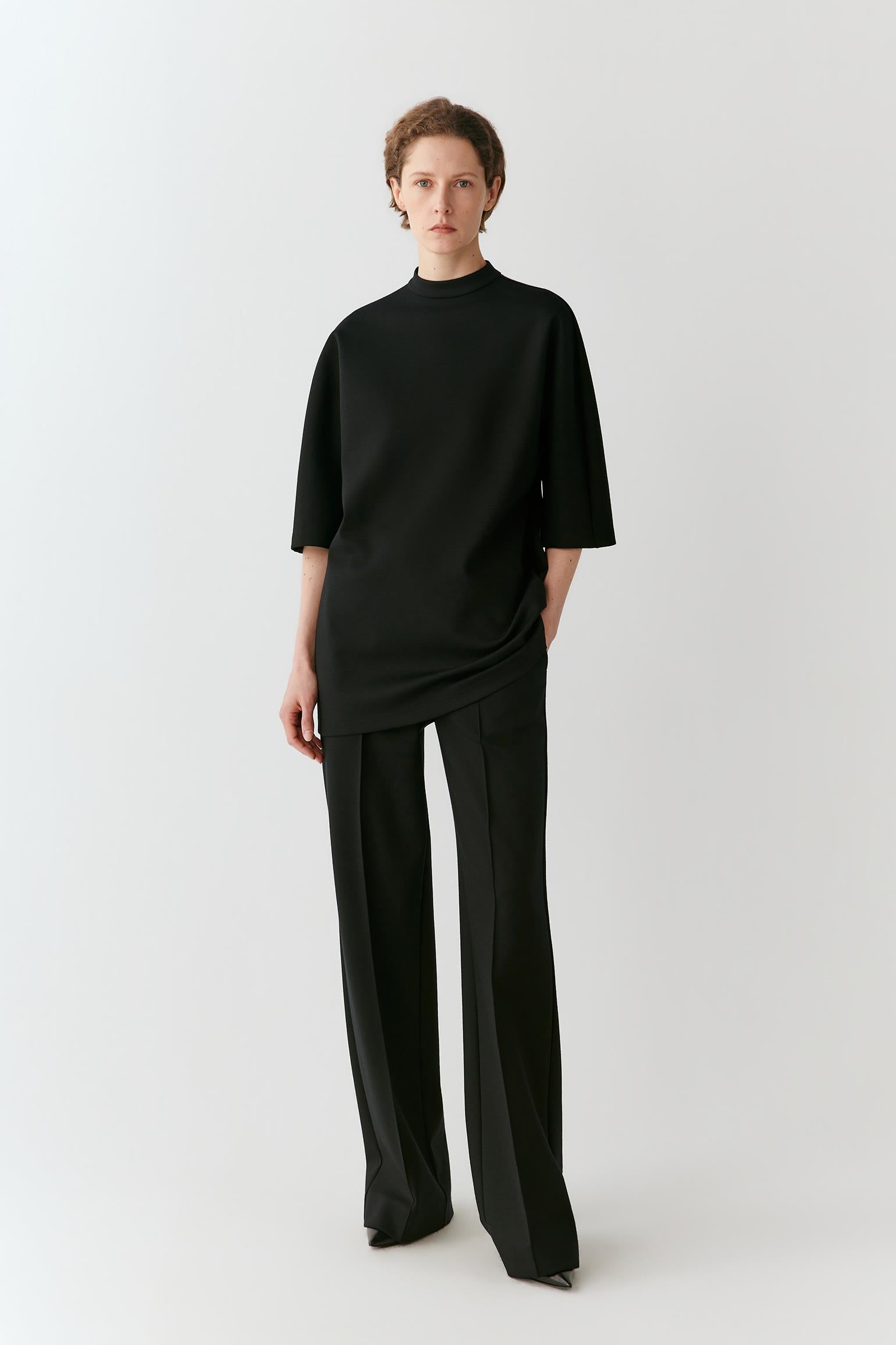 A model stands against a light gray studio background wearing a black asymmetrical mock-neck top and matching black wide-leg trousers.