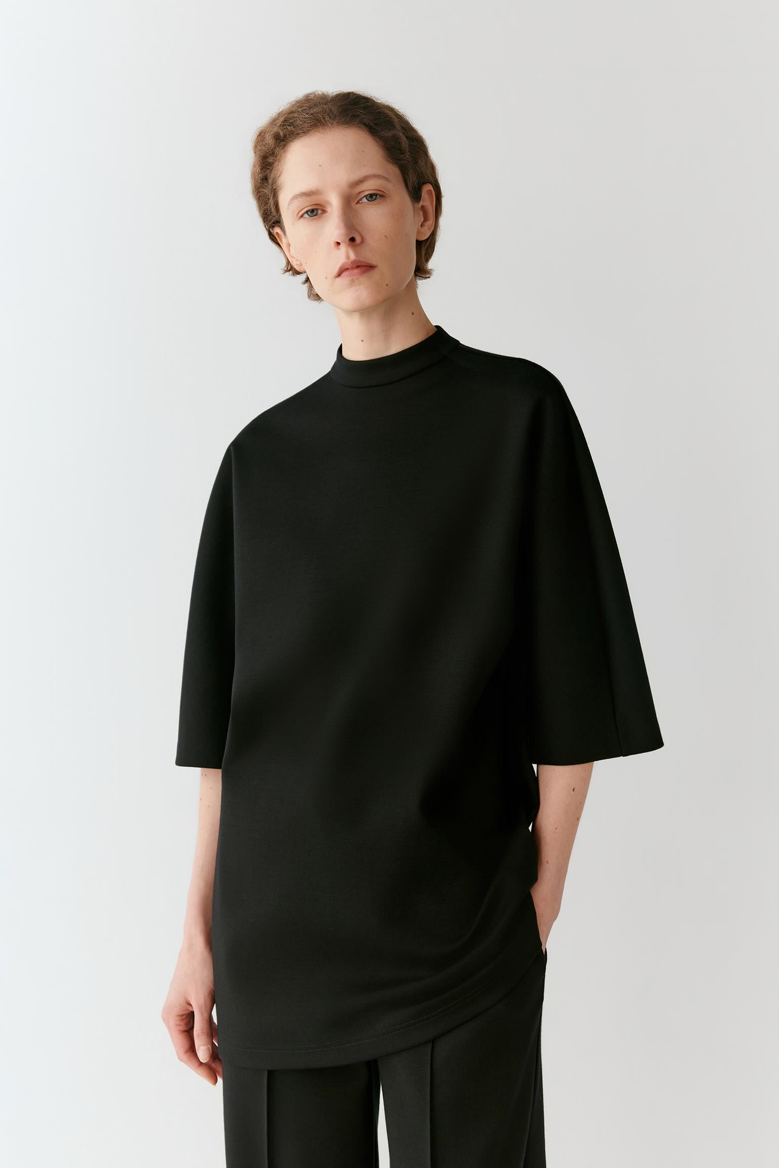 A model wearing a black oversized mock-neck t-shirt and matching black trousers, standing against a light gray studio background.