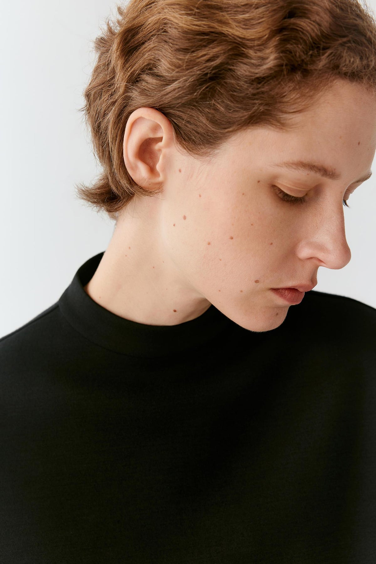Close-up of a person with short auburn hair wearing a black mock neck top against a light gray background.