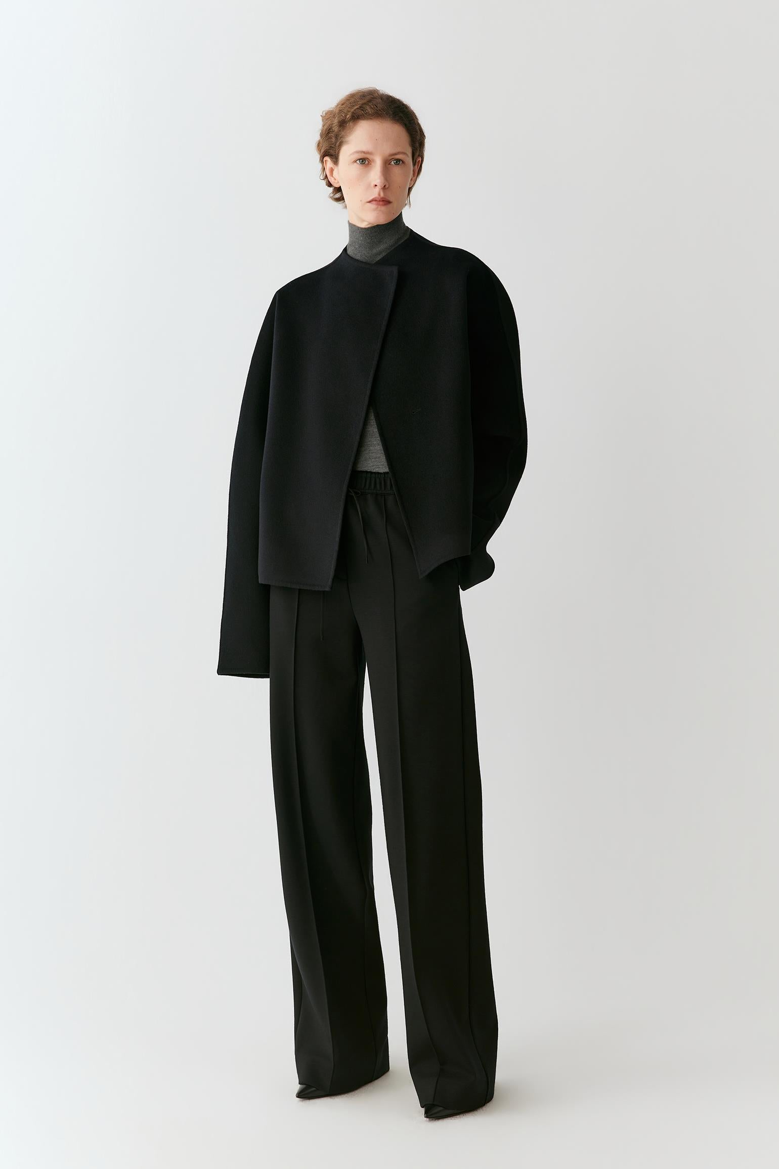A model stands against a light gray studio background wearing a black asymmetrical draped jacket, black wide-leg trousers, and a dark gray mock-neck top.