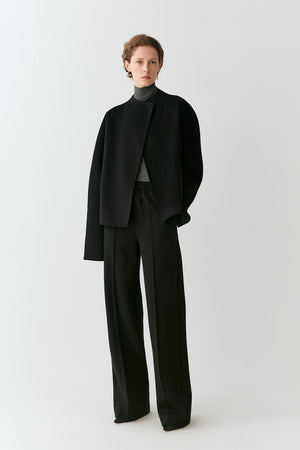 A model stands against a light gray studio background wearing a black asymmetrical draped jacket, black wide-leg trousers, and a dark gray mock-neck top.