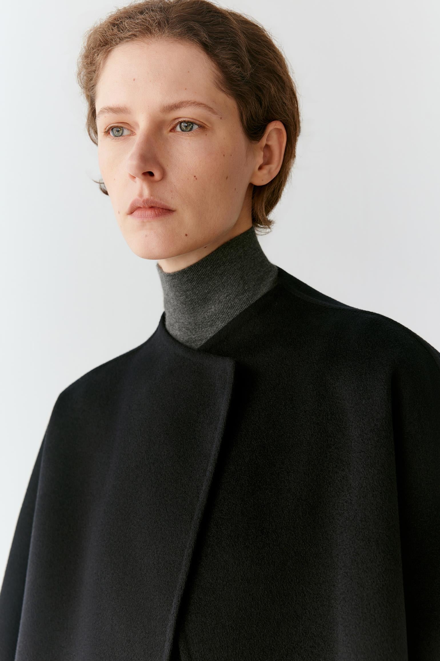A close-up portrait of a model wearing a black cape-style coat over a dark gray mock neck sweater, set against a plain light gray background.