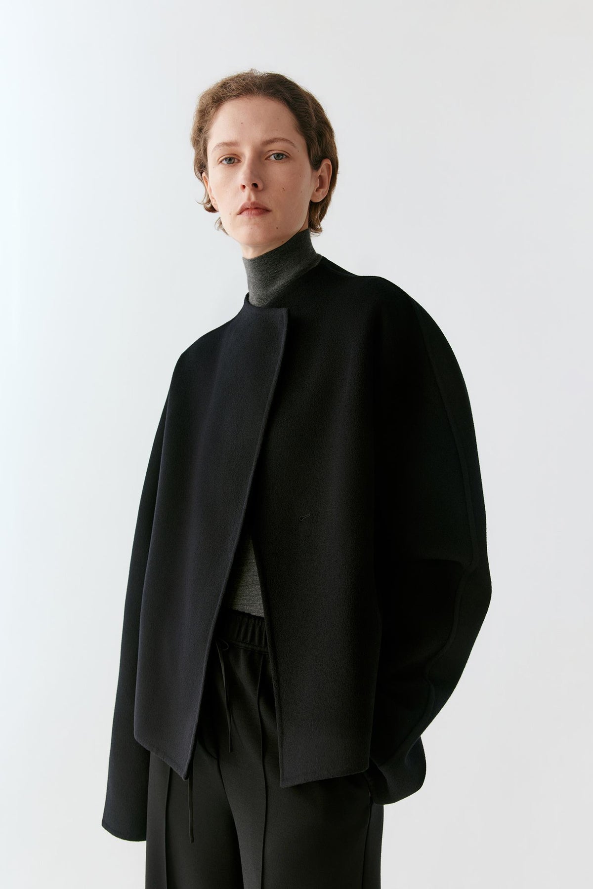 A model wearing a black asymmetrical draped cape jacket over a black turtleneck sweater and black trousers, photographed against a clean white studio background.