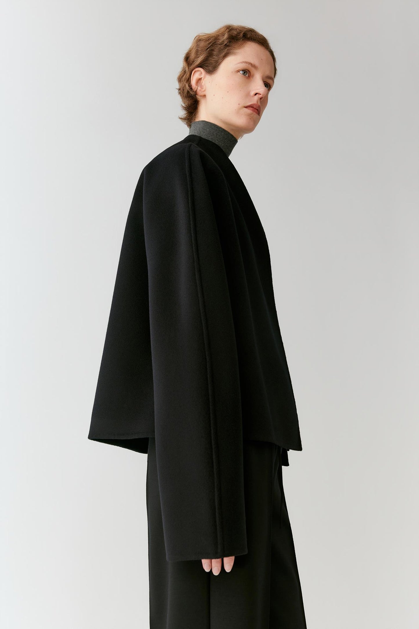 A model wearing a black high-neck, oversized cape-style jacket with wide draped sleeves and matching black trousers, photographed against a neutral light gray studio background.