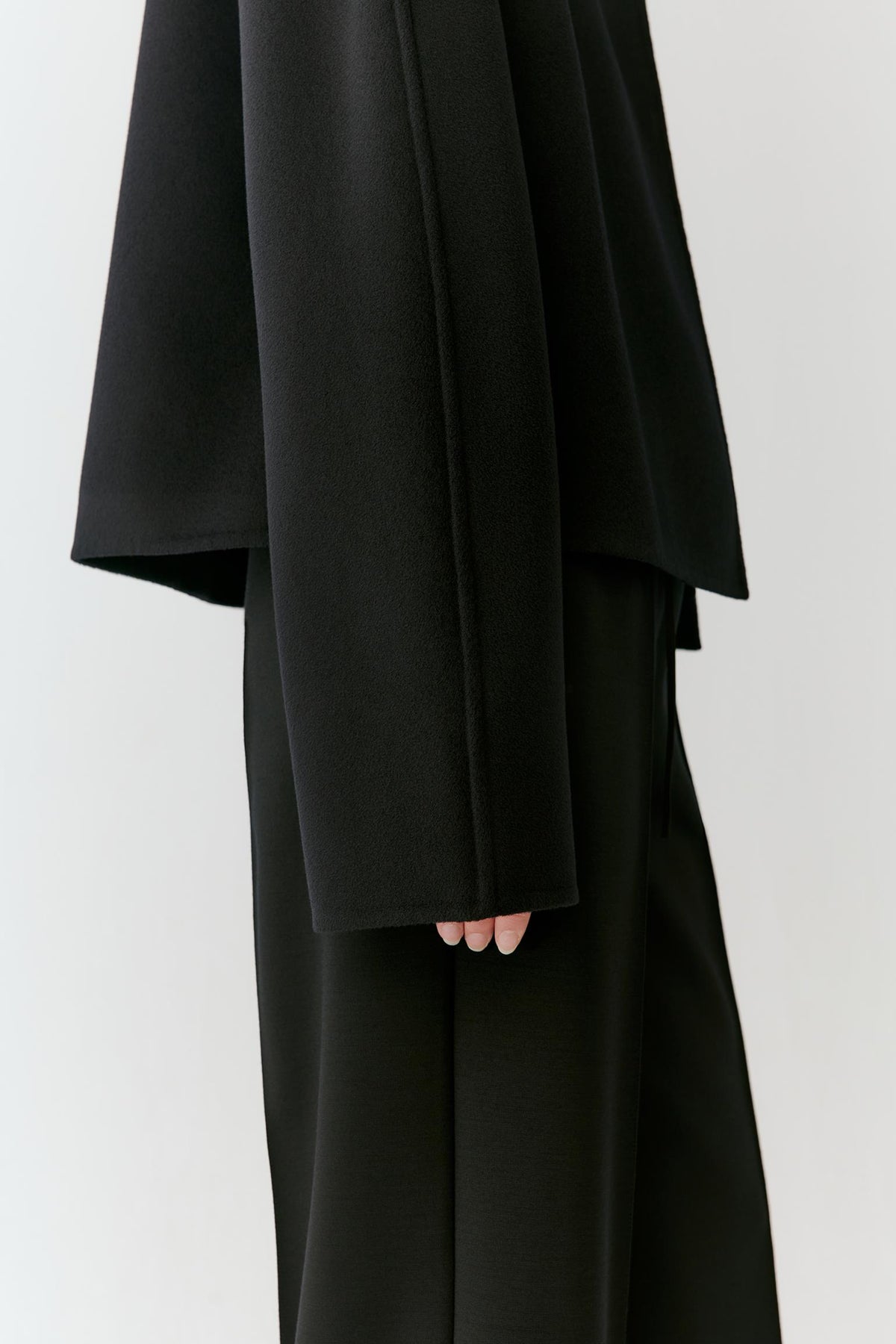 A close-up of a person wearing a black draped, oversized long-sleeve top or dress with matching black wide-leg bottoms, shown against a plain light gray background.