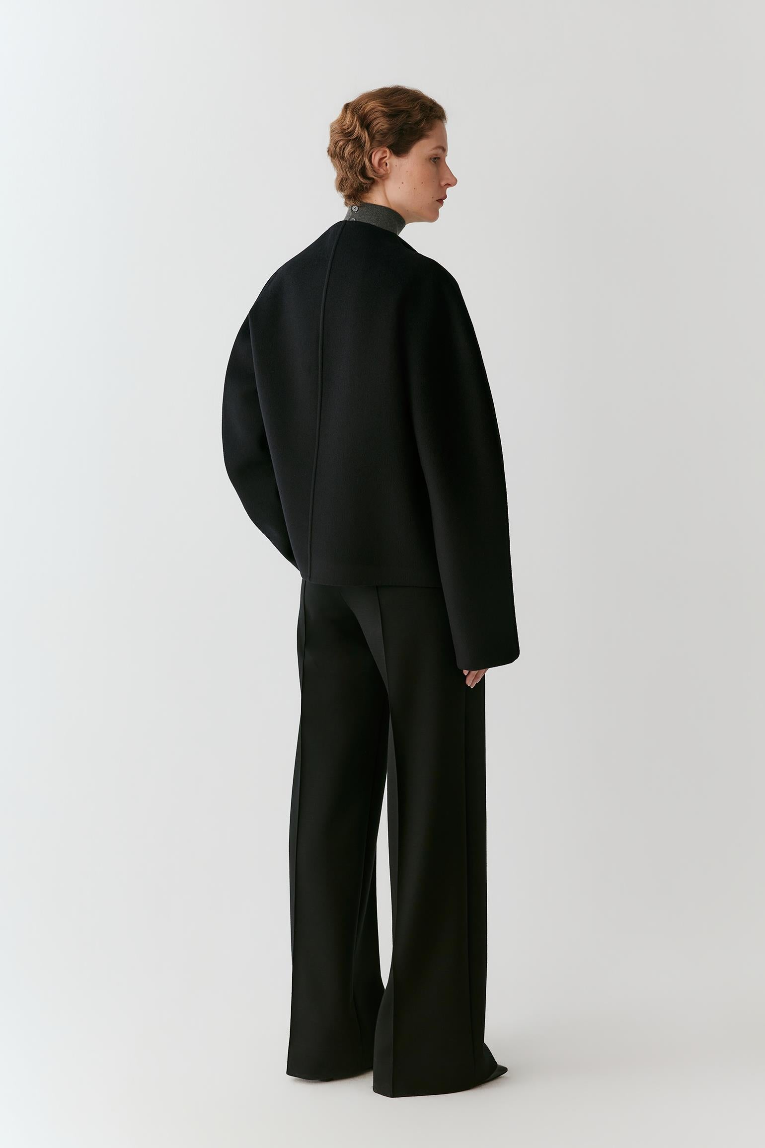 A model shown from the back wearing a black long-sleeve cropped jacket and matching wide-leg trousers against a light gray studio background.