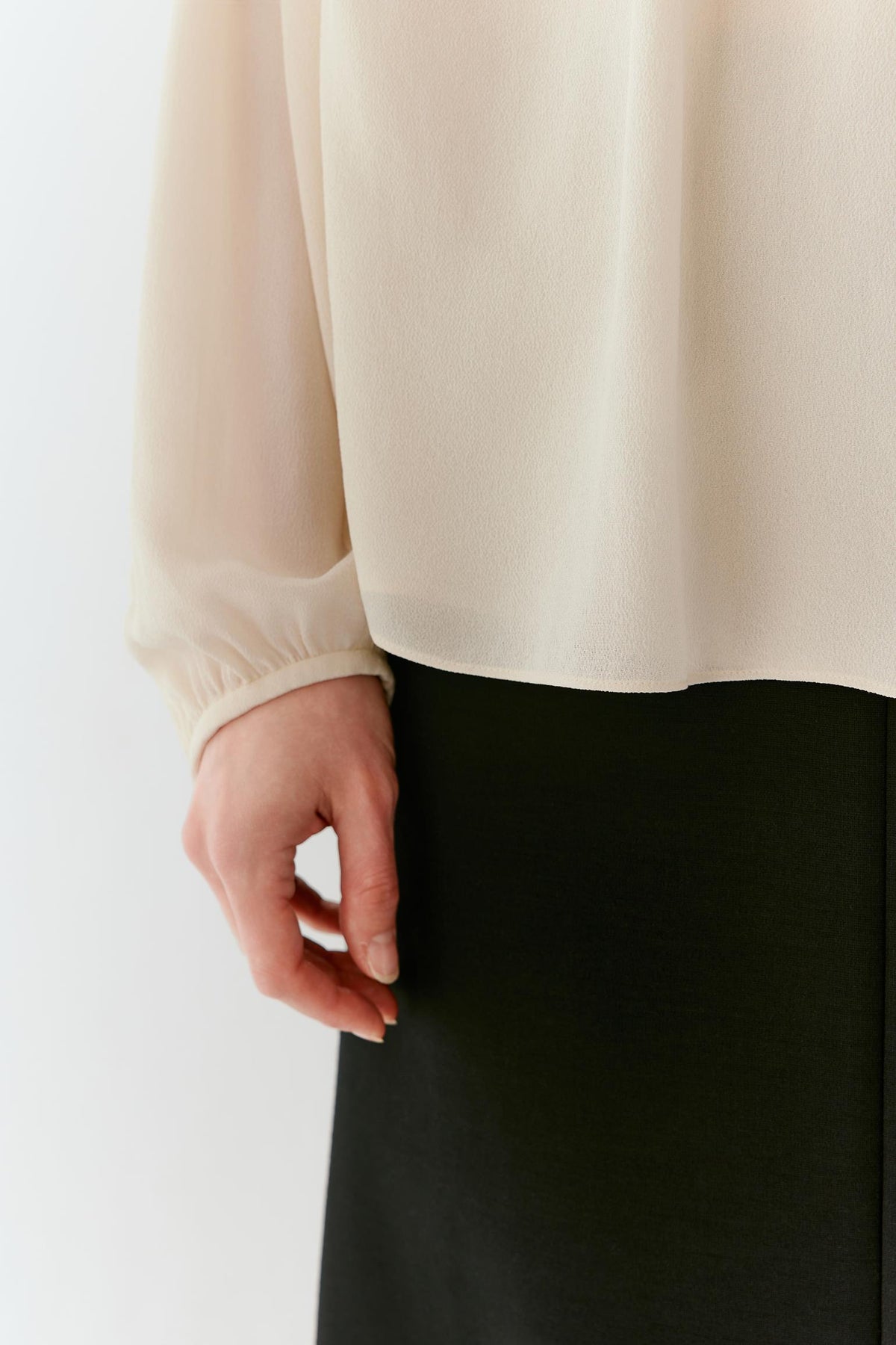 Close-up of a person wearing a cream-colored long-sleeve blouse and a black skirt, with the hand resting near the waistline against a plain white background.