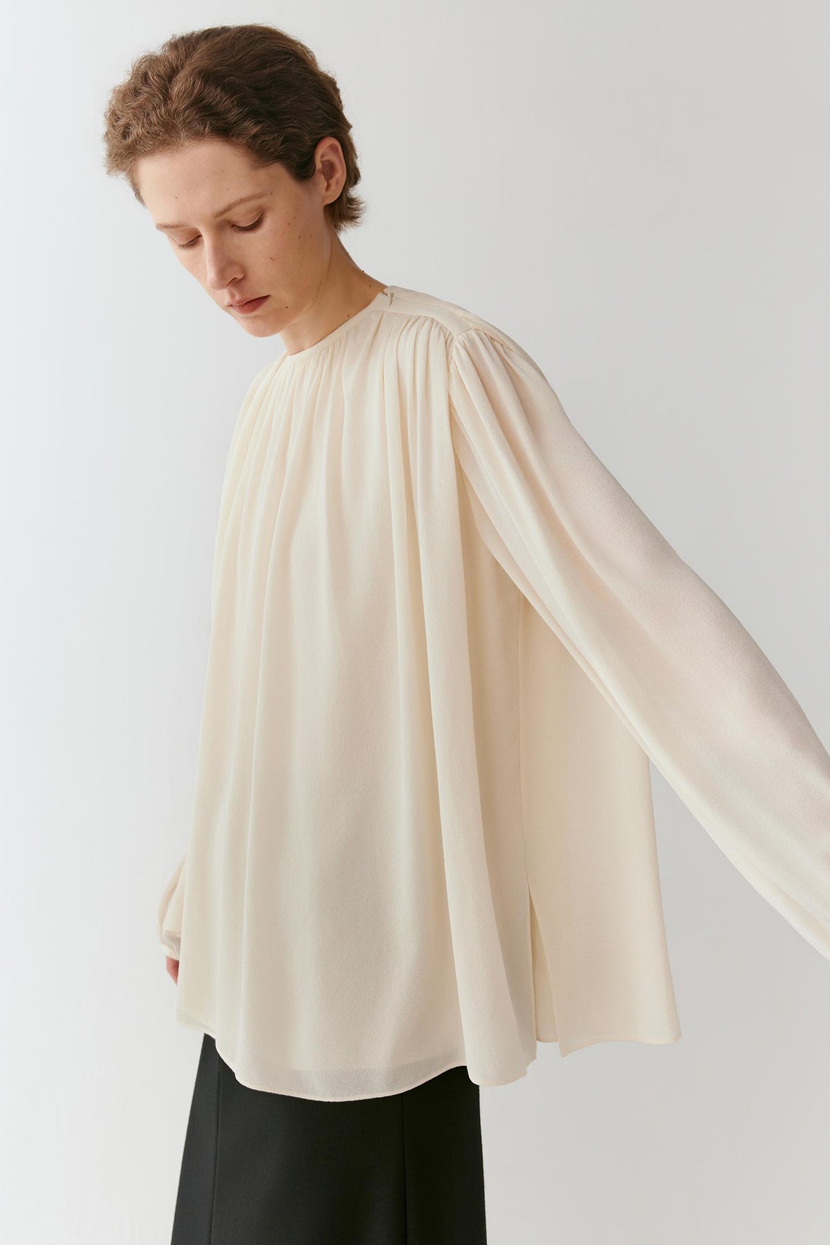 A woman wearing a cream-colored, semi-sheer long-sleeve blouse with a high gathered neckline and black trousers, photographed against a light gray studio background.