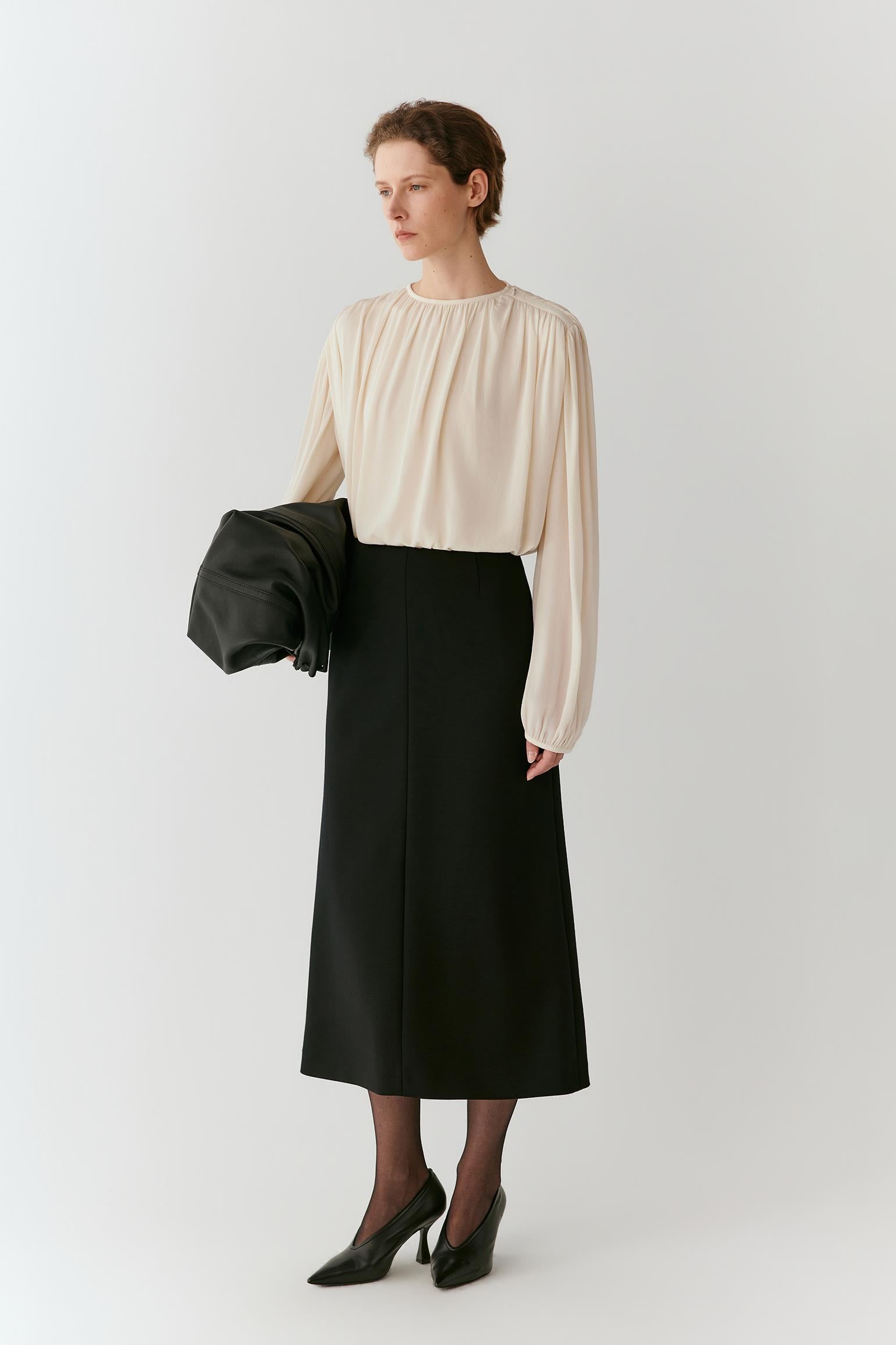 A woman stands against a light gray studio background wearing a cream long-sleeve blouse with a gathered neckline and a black midi skirt. She holds a black clutch in one hand and wears black pointed-toe heels.