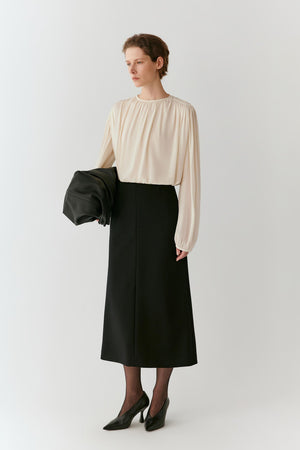 A woman stands against a light gray studio background wearing a cream long-sleeve blouse with a gathered neckline and a black midi skirt. She holds a black clutch in one hand and wears black pointed-toe heels.
