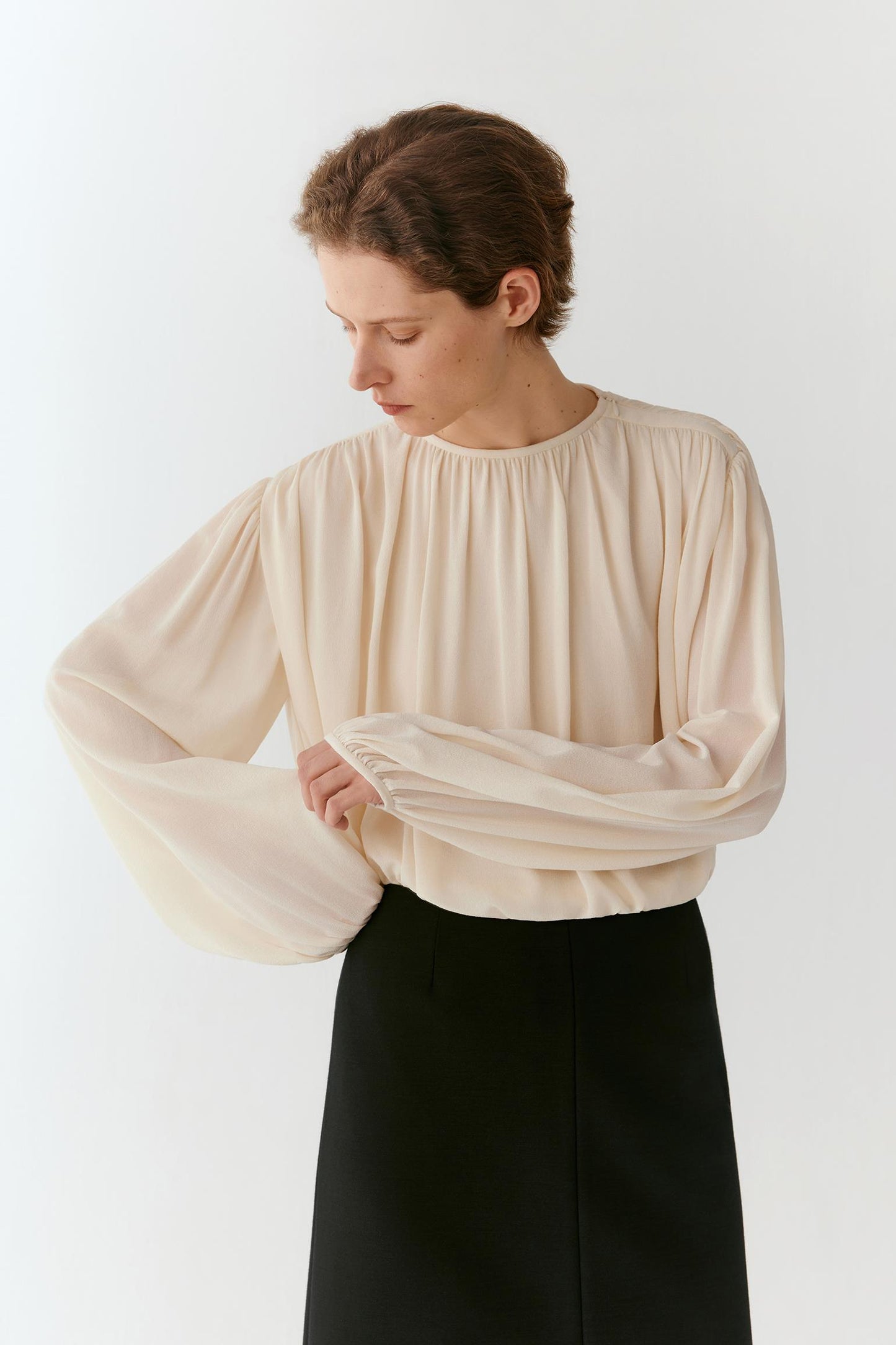 A woman wearing a cream-colored long-sleeve blouse with dramatic balloon sleeves and a gathered neckline, paired with a black skirt, photographed against a plain white studio background.