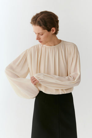 A woman wearing a cream-colored long-sleeve blouse with dramatic balloon sleeves and a gathered neckline, paired with a black skirt, photographed against a plain white studio background.