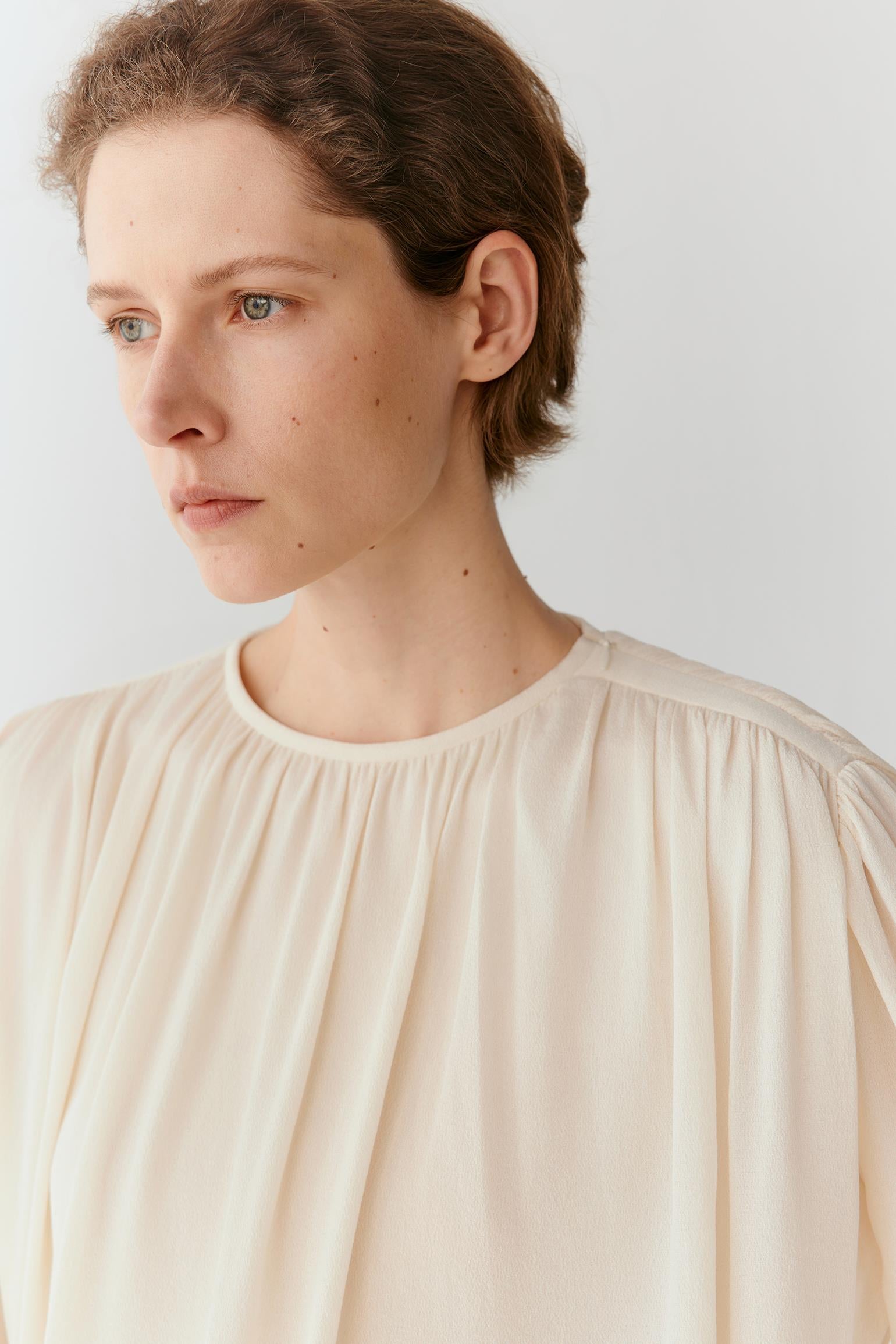 A close-up shot of a model wearing a cream-colored, lightweight pleated blouse against a plain light gray background.