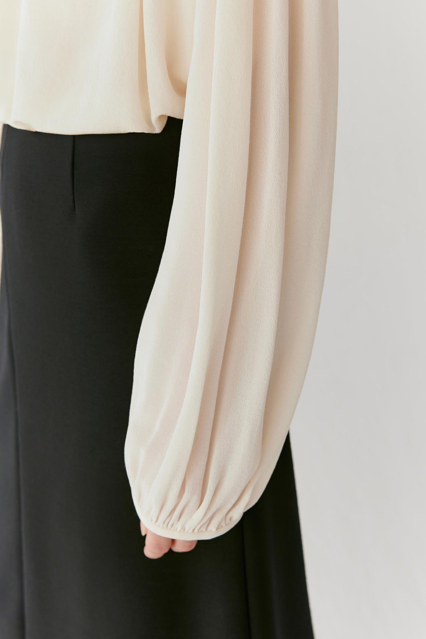 Close-up of a model wearing a cream-colored blouse with dramatic balloon sleeves and black trousers, against a plain light gray background.