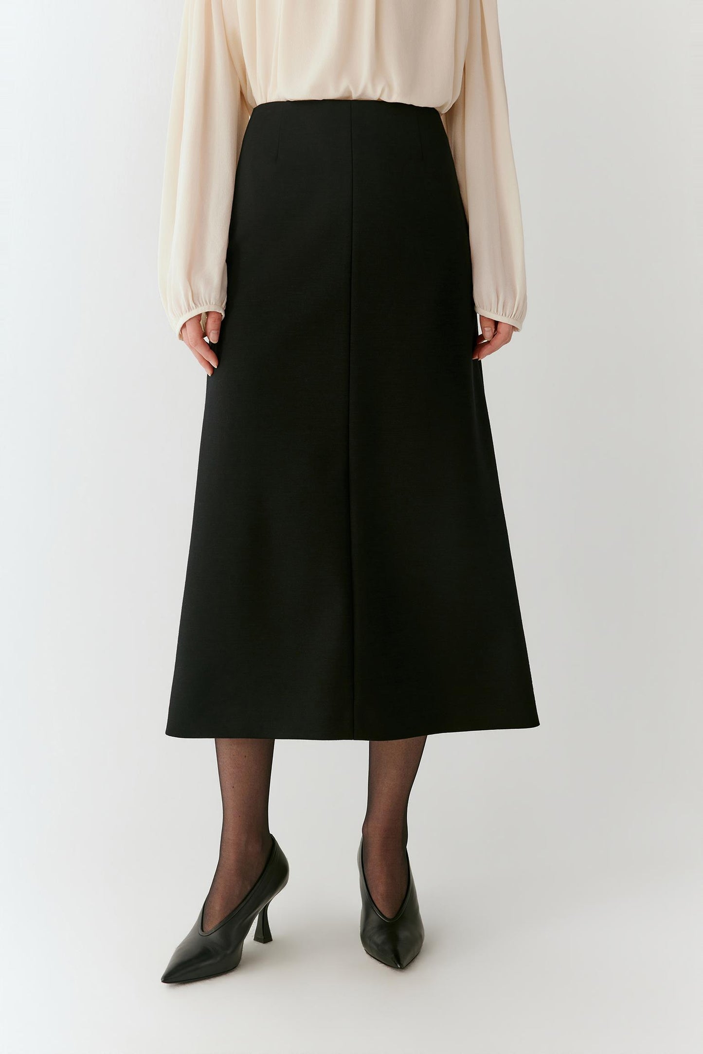 A model is shown from the waist down wearing a black midi skirt, a cream blouse, sheer black tights, and black pointed-toe heels, standing against a plain light gray studio background.