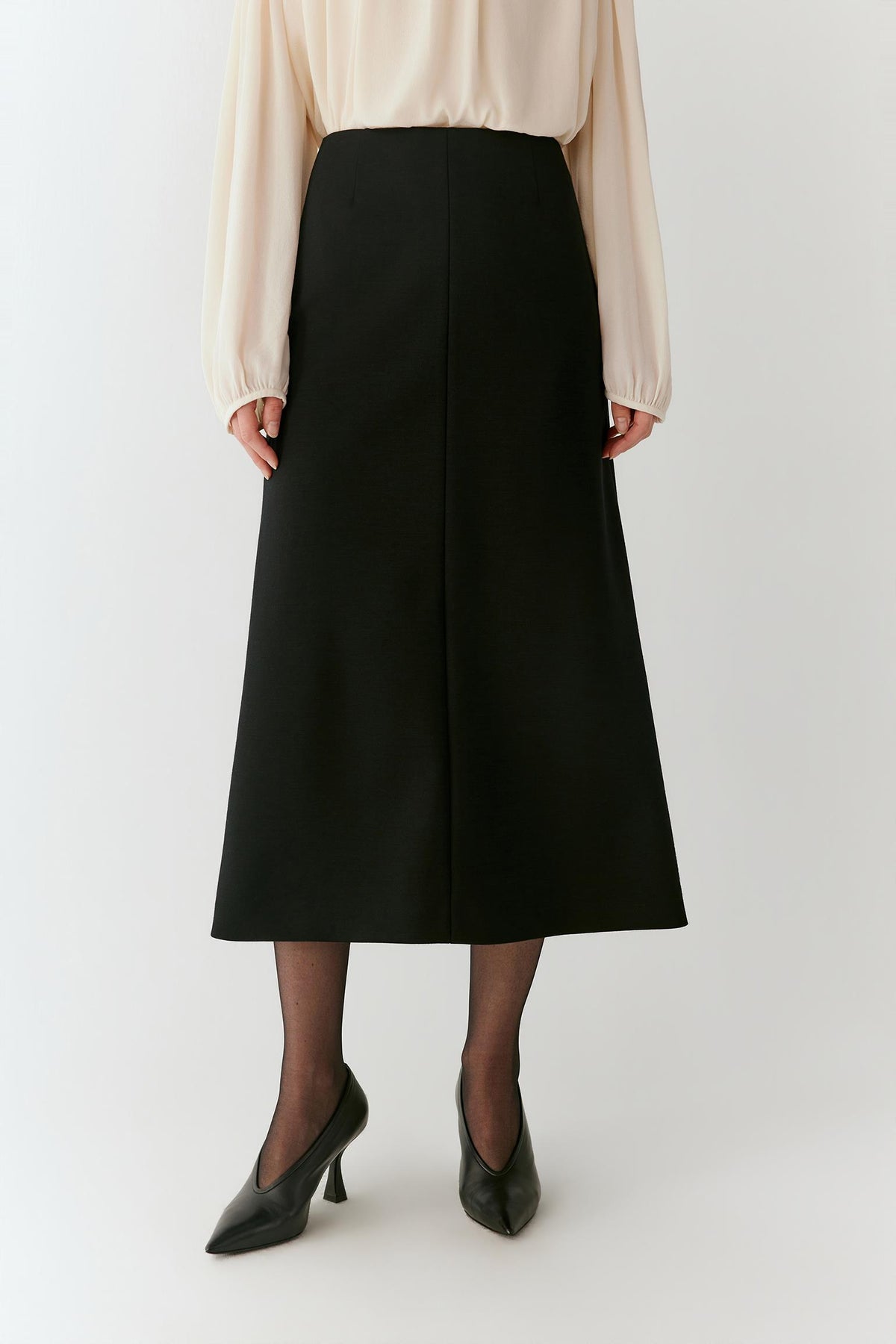 A model is shown from the waist down wearing a black midi skirt, a cream blouse, sheer black tights, and black pointed-toe heels, standing against a plain light gray studio background.