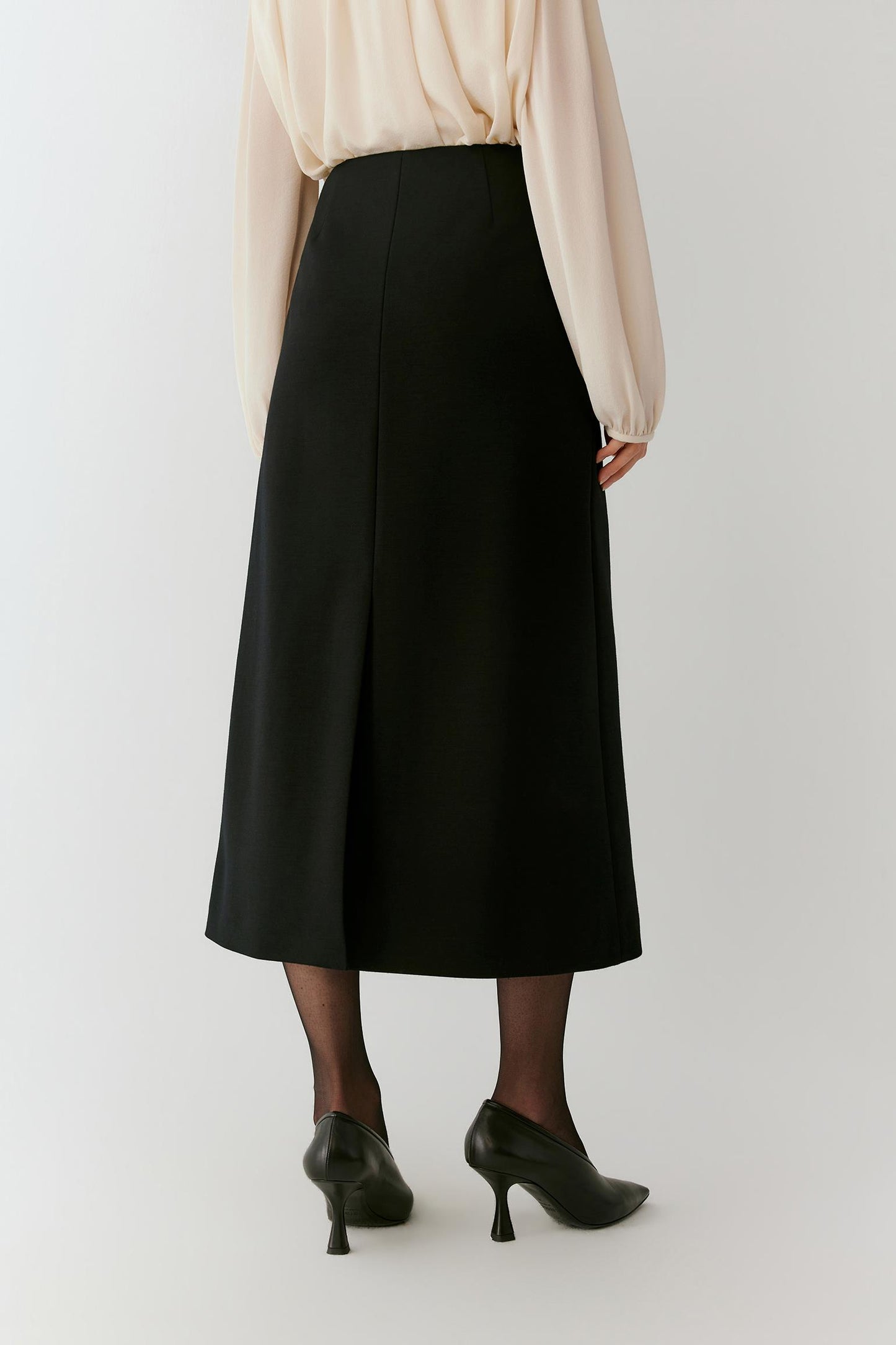 A model shown from the back wearing a black midi skirt, a cream long-sleeve blouse, sheer black tights, and black pointed-toe heels against a light gray studio background.
