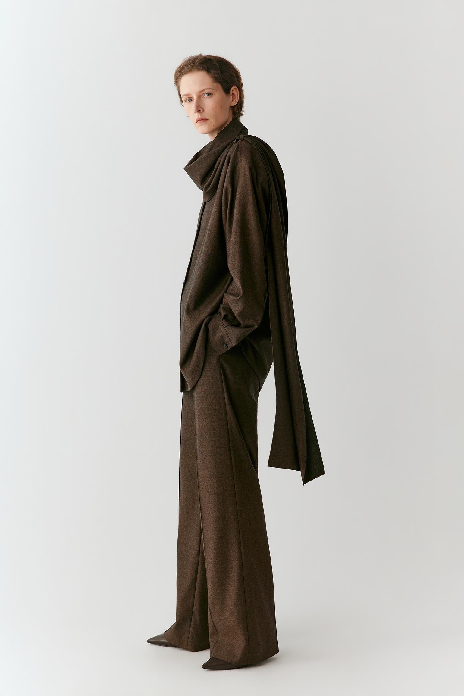 A model wearing a dark brown, draped, wide-sleeved outfit with matching wide-leg trousers, standing in profile against a light gray studio background.