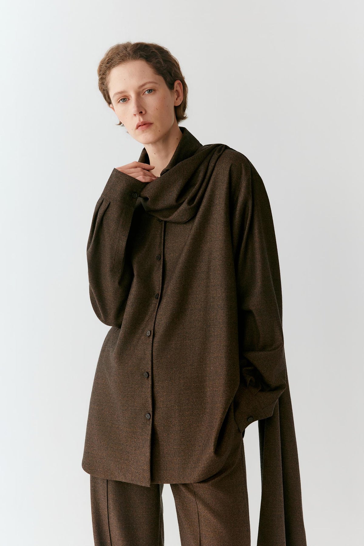 A model wearing a dark brown, oversized long-sleeve button-up shirt with a draped, high collar and matching brown trousers, photographed against a light gray studio background.