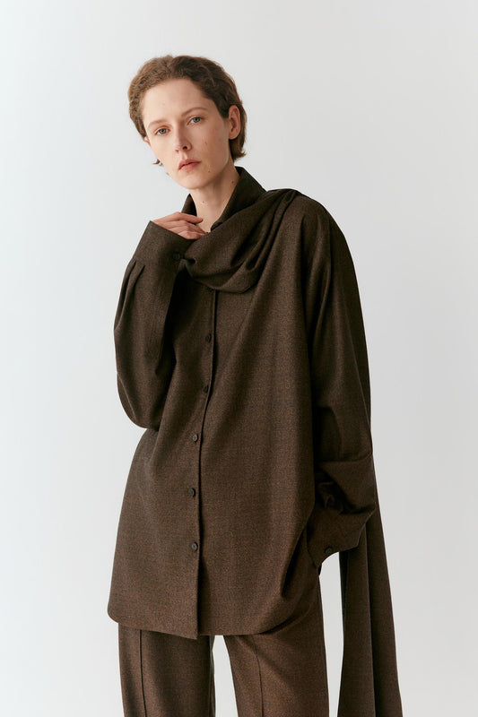 A model wearing a dark brown, oversized long-sleeve button-up shirt with a draped, high collar and matching brown trousers, photographed against a light gray studio background.