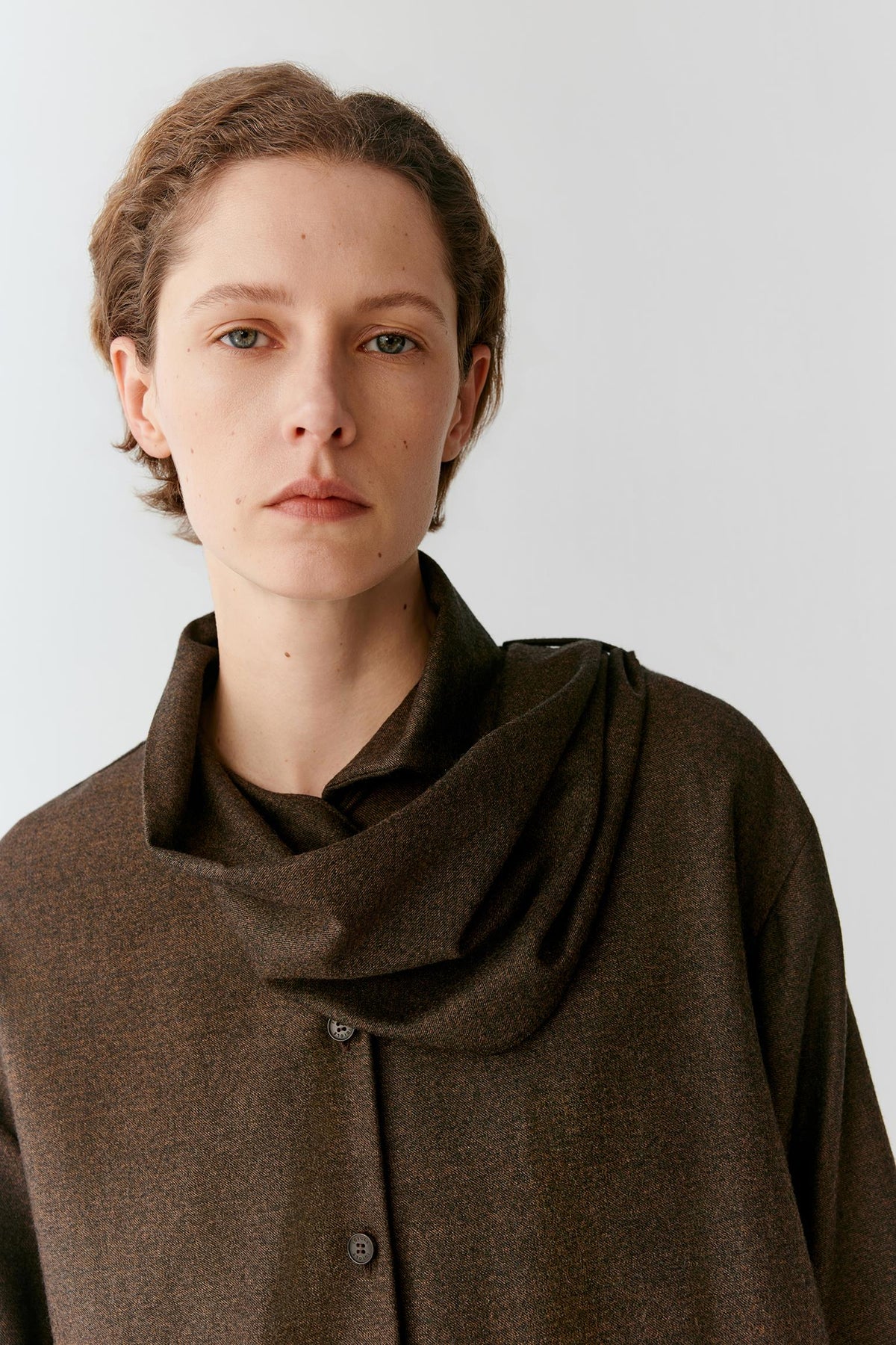 A model with short brown hair wearing a dark brown textured button-up shirt with a draped, high folded collar, photographed against a light gray studio background.