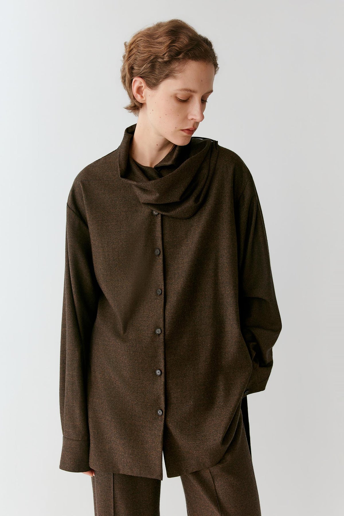 A woman wearing an oversized dark brown cowl-neck button-front top with matching pants, photographed against a plain light gray studio background.