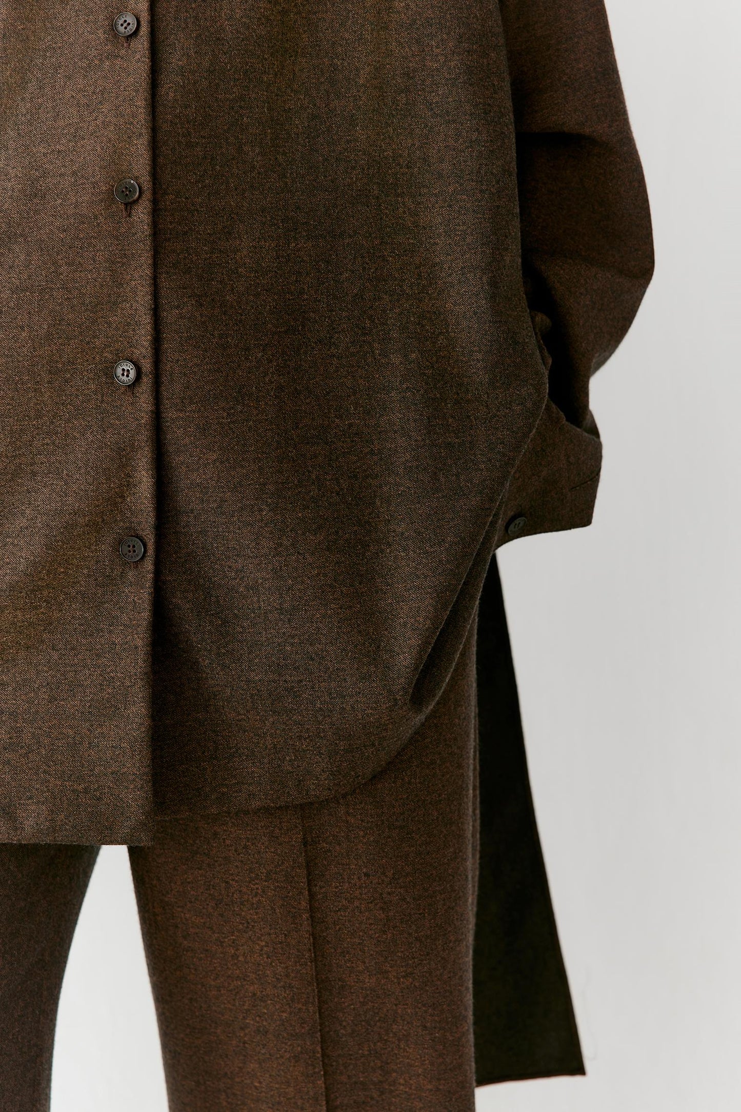 Close-up of a brown textured button-front jacket or overshirt with matching brown trousers, shown on a mannequin against a light gray studio background.