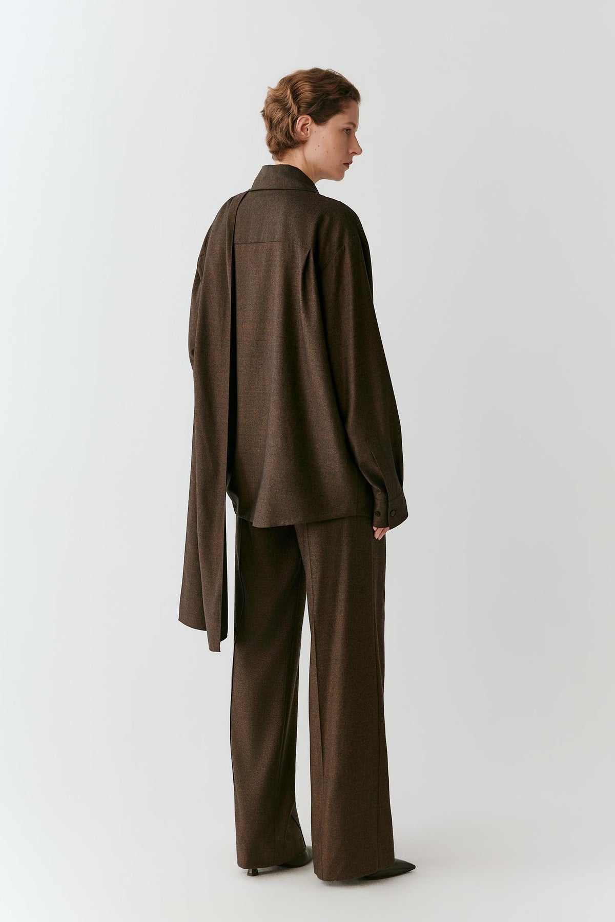 A model shown from the back wearing a dark brown oversized long-sleeve shirt and matching wide-leg trousers against a light gray studio background.
