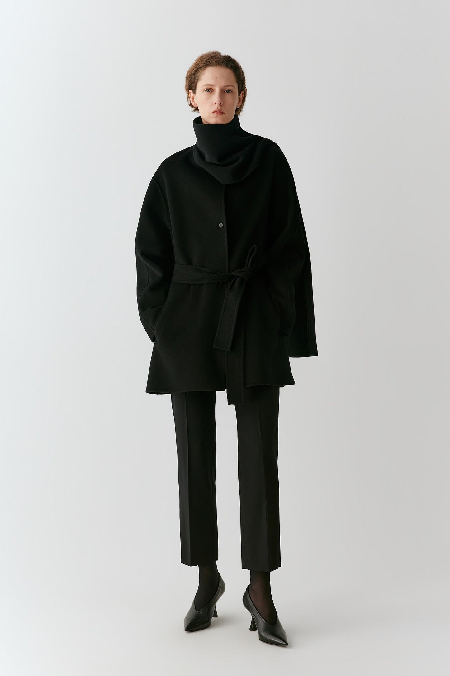A model stands against a light gray studio background wearing a black cowl-neck belted coat, black trousers, and black pointed shoes.