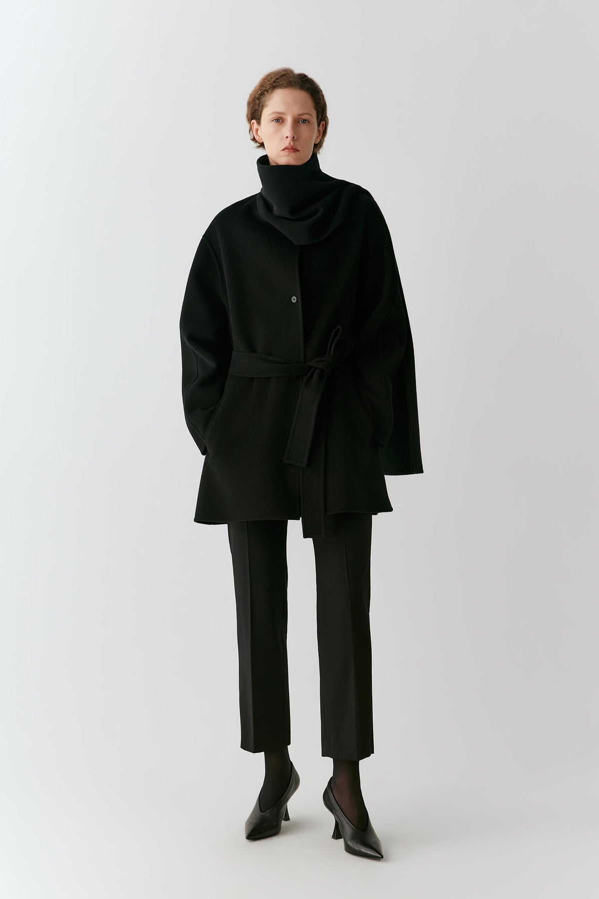 A model stands against a light gray studio background wearing a black cowl-neck belted coat, black trousers, and black pointed shoes.