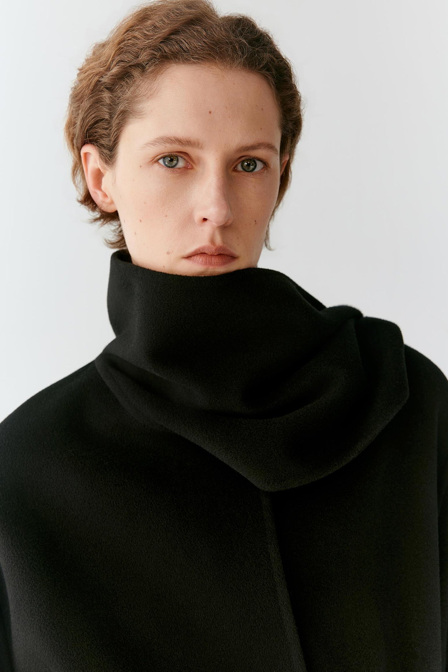 A close-up portrait of a model wearing a black high-neck cowl neck sweater against a light gray background.