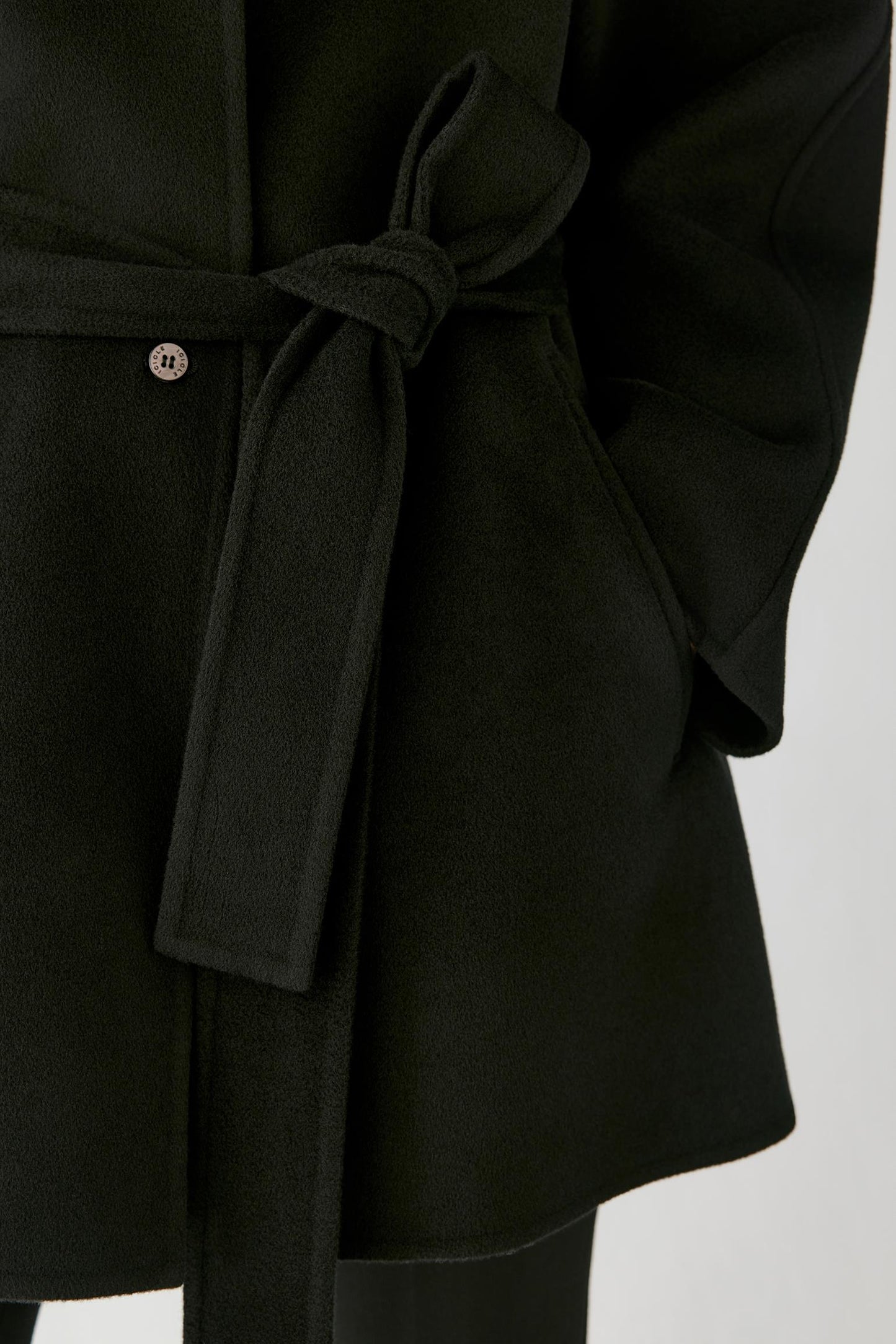 Close-up of a black belted coat with a matching tie belt and a small metallic button detail, shown against a light gray background.