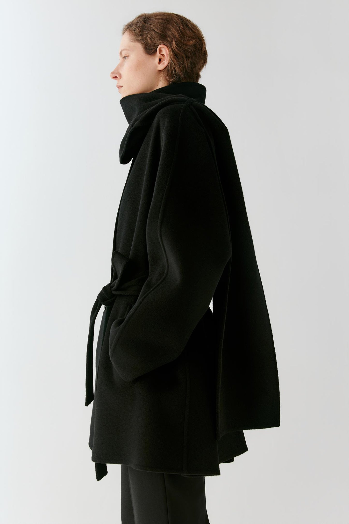 A model shown in profile wearing a black belted coat with a high structured collar, photographed against a clean light gray studio background.