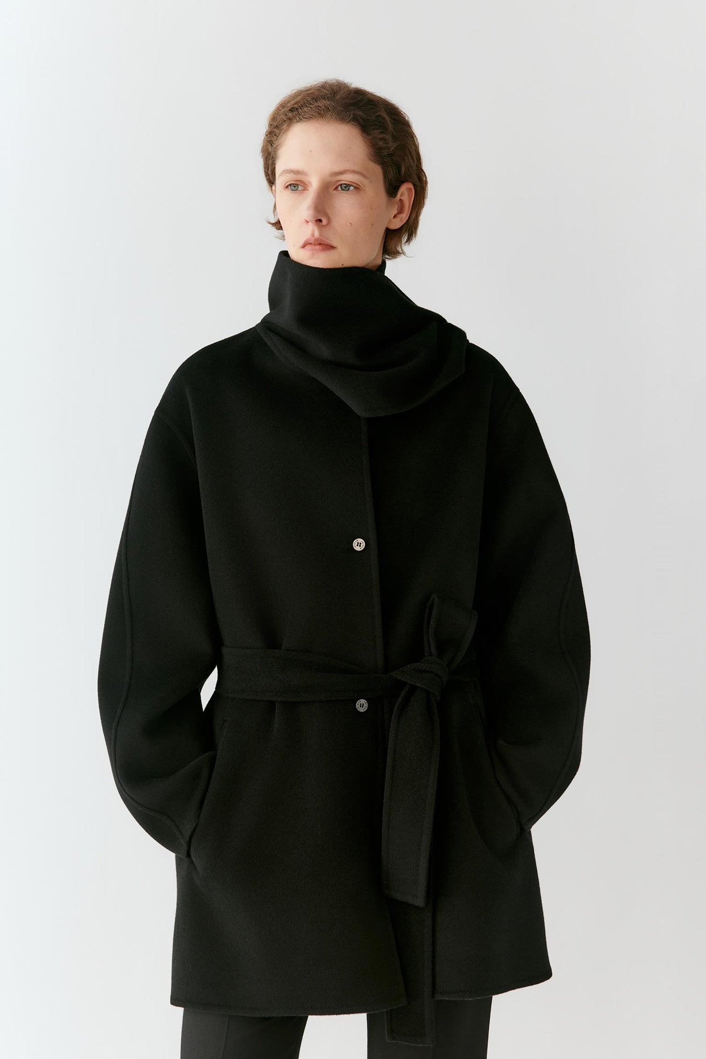 A model wearing a black wrap-style coat with a high folded collar, front buttons, and a self-tie belt, photographed against a plain light gray studio background.