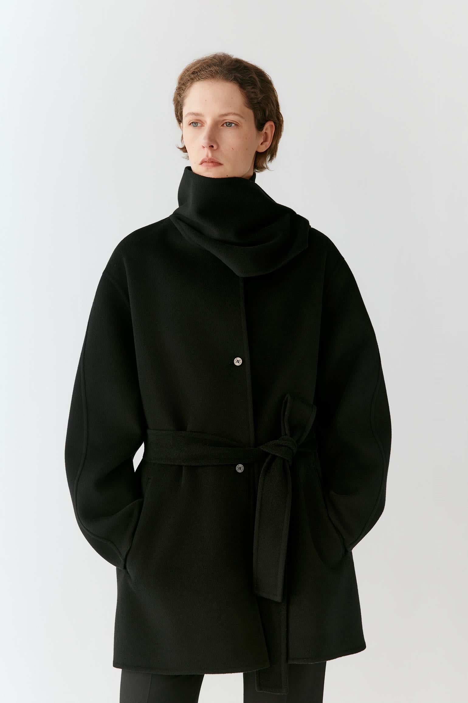 A model wearing a black wrap-style coat with a high folded collar, front buttons, and a self-tie belt, photographed against a plain light gray studio background.