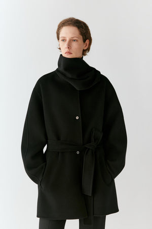 A model wearing a black wrap-style coat with a high folded collar, front buttons, and a self-tie belt, photographed against a plain light gray studio background.