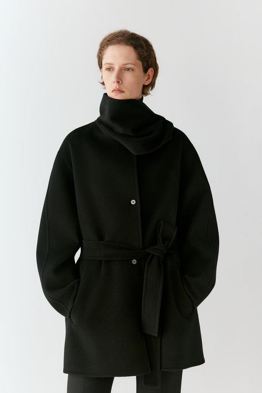A model wearing a black wrap-style coat with a high folded collar, front buttons, and a self-tie belt, photographed against a plain light gray studio background.