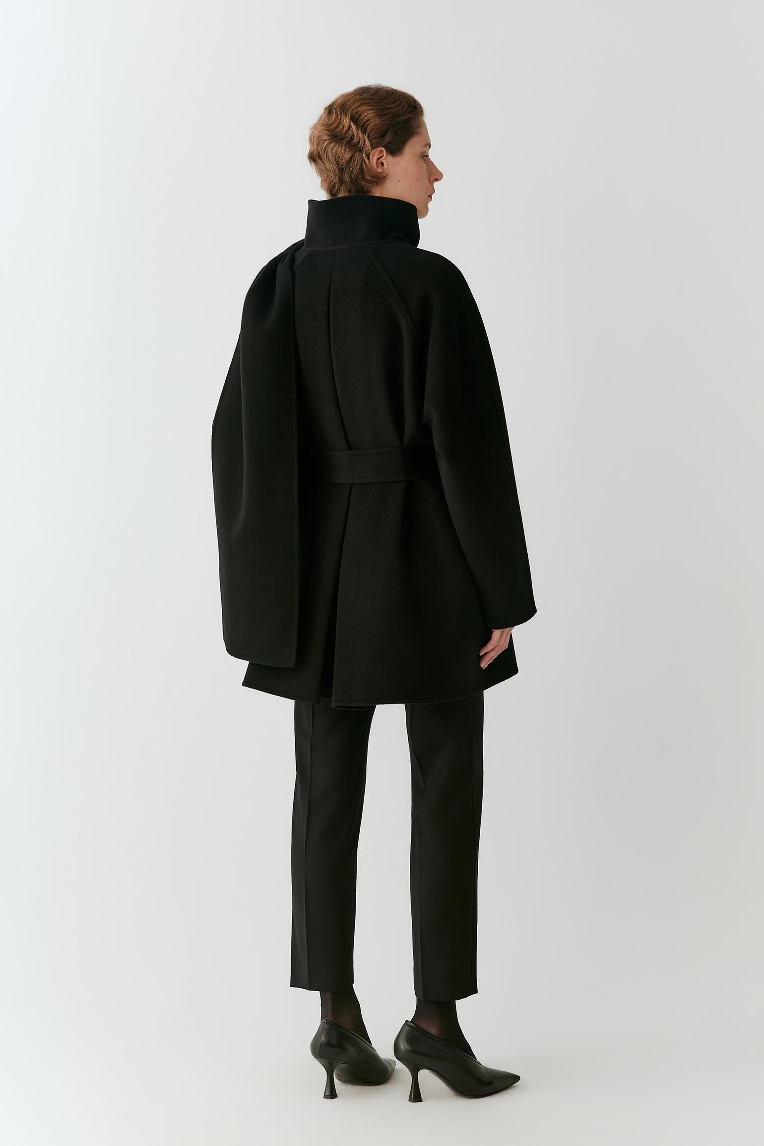 A model shown from the back wearing a black high-neck belted coat, black trousers, and black pointed-toe heels against a light gray studio background.