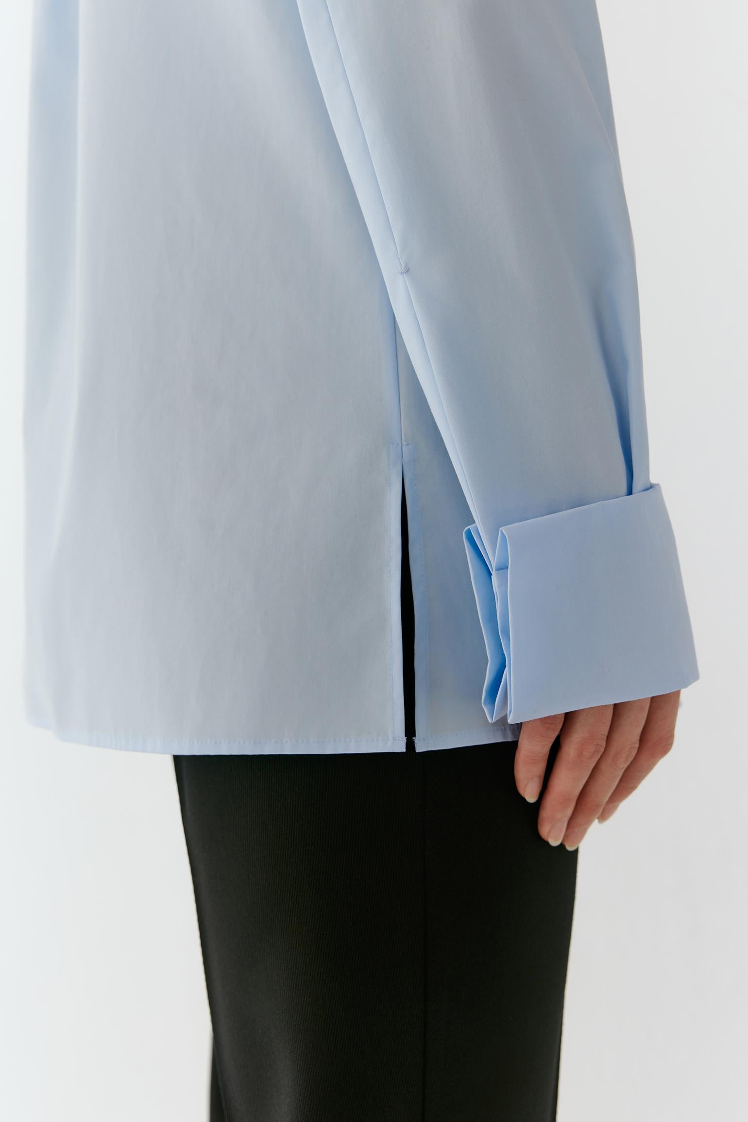 Close-up of a person wearing a light blue long-sleeve shirt with a wide cuff and black trousers against a plain light background.