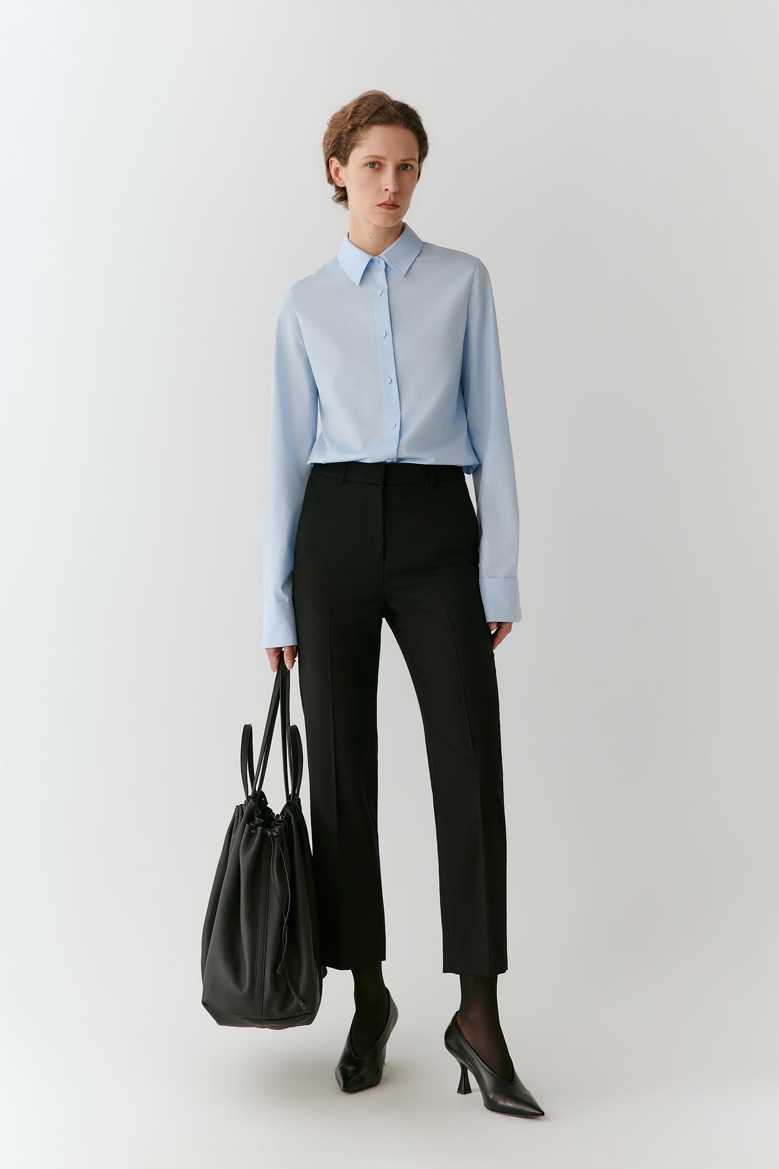 A woman stands against a light gray studio background wearing a light blue long-sleeve button-up shirt, black cropped trousers, black pointed heels, and carrying a black tote bag.