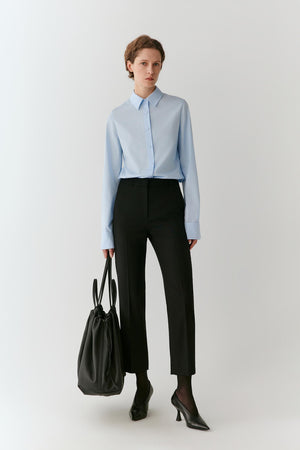A woman stands against a light gray studio background wearing a light blue long-sleeve button-up shirt, black cropped trousers, black pointed heels, and carrying a black tote bag.