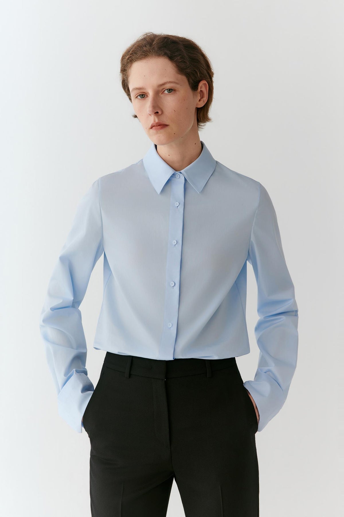 A model wearing a light blue long-sleeve button-down shirt tucked into black trousers, standing against a plain light gray studio background.