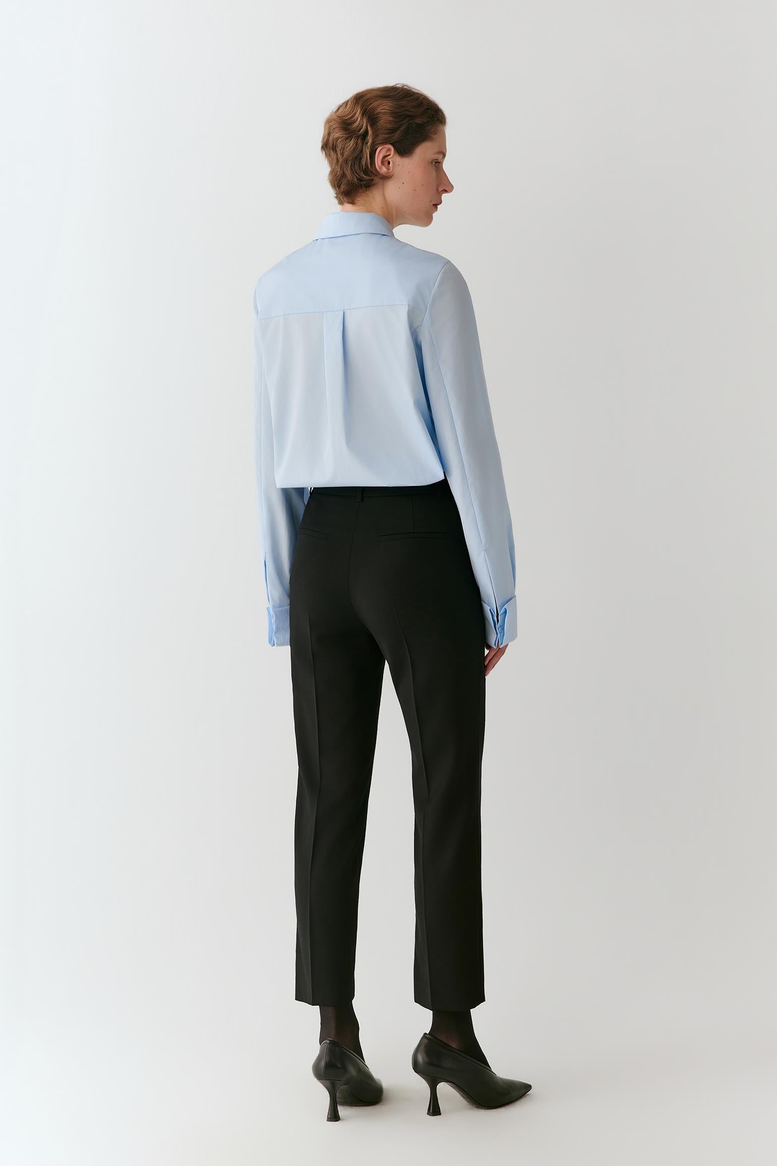 A woman shown from the back wearing a light blue long-sleeve button-up shirt, black cropped trousers, and black pointed-toe heels against a plain light gray studio background.