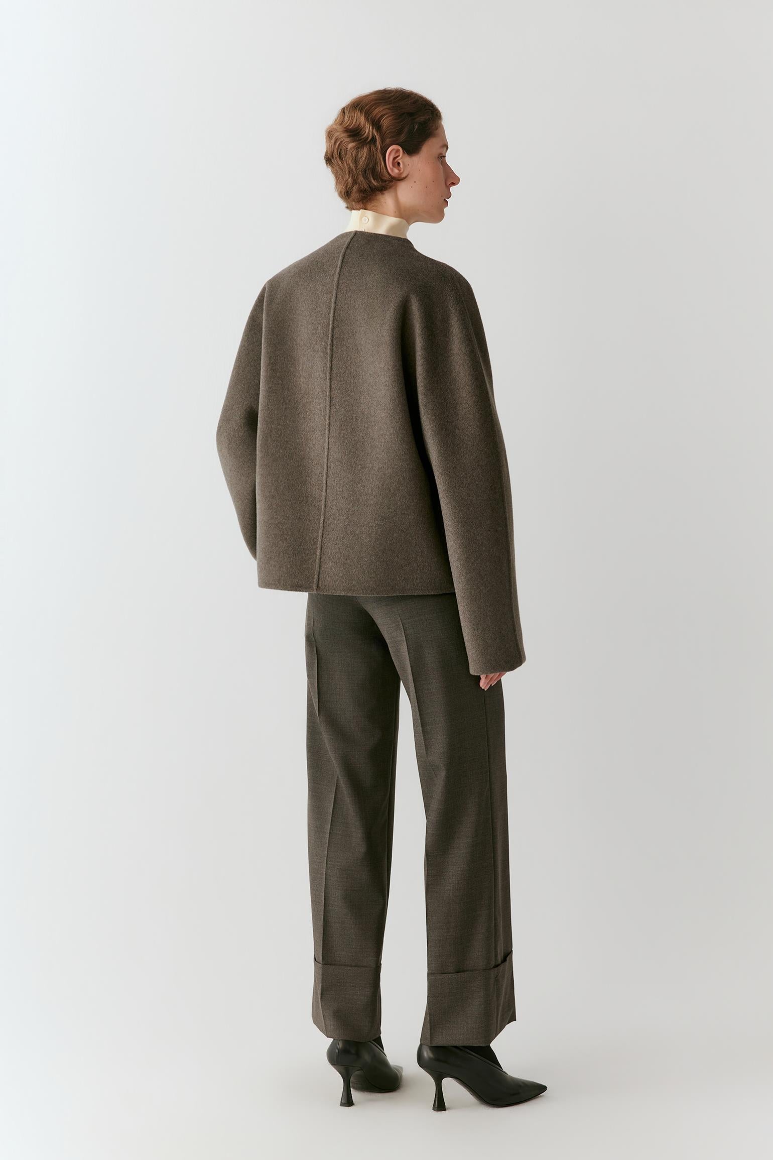 A model shown from the back wearing a dark brown cropped jacket and matching wide-leg trousers with black pointed-toe heels, set against a plain light gray studio background.