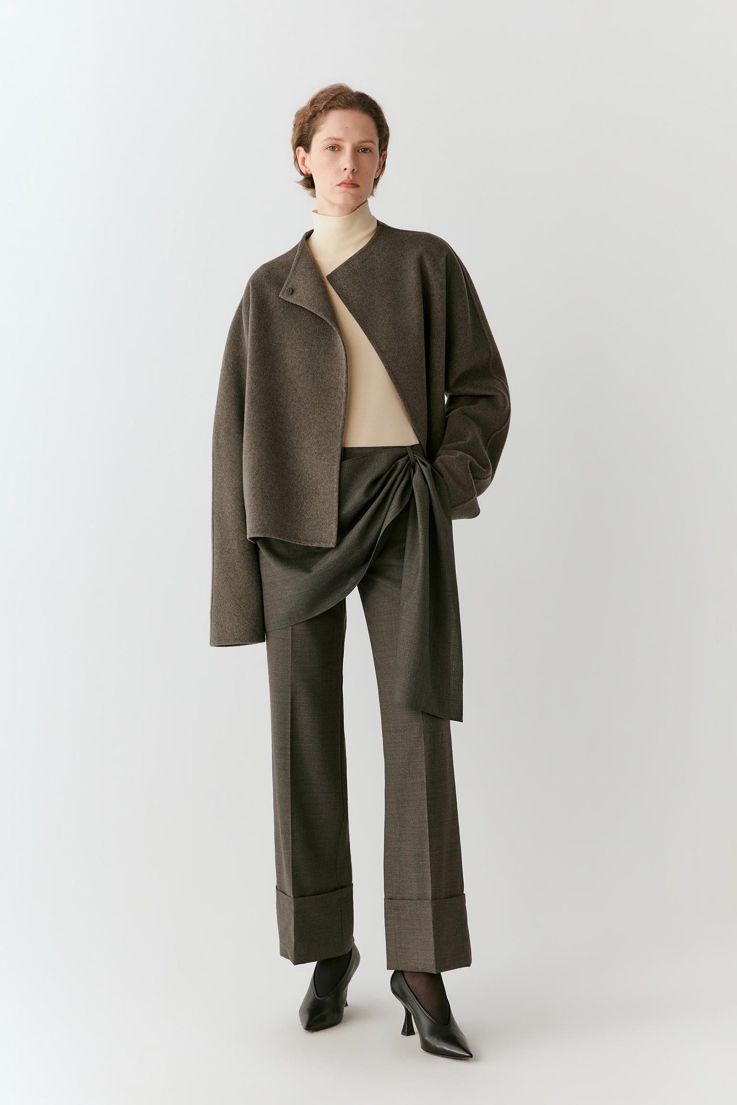 A model stands against a light gray studio background wearing a dark brown asymmetrical wrap jacket, matching dark brown wide-leg trousers, a cream mock-neck top, and black pointed-toe heels.
