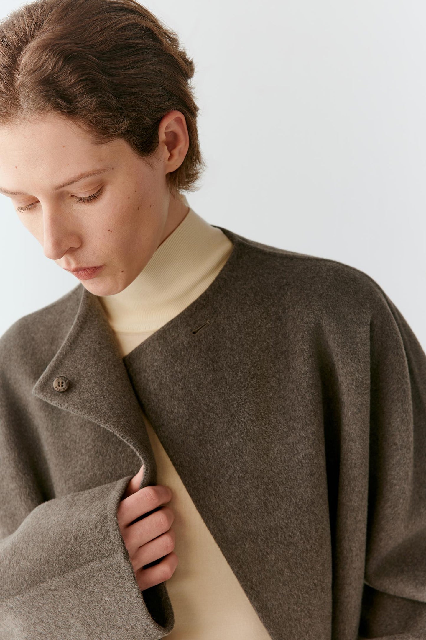 A close-up of a model wearing a dark brown wool-blend coat over a cream turtleneck sweater, set against a plain light gray background.