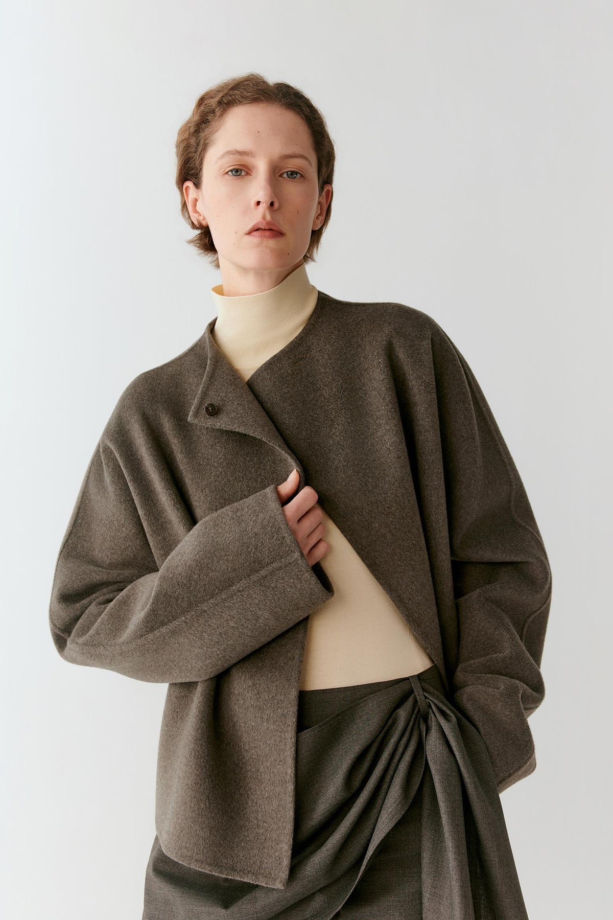 A model wearing a dark brown textured wrap jacket over a cream mock neck top and a dark gray skirt, photographed against a light gray studio background.