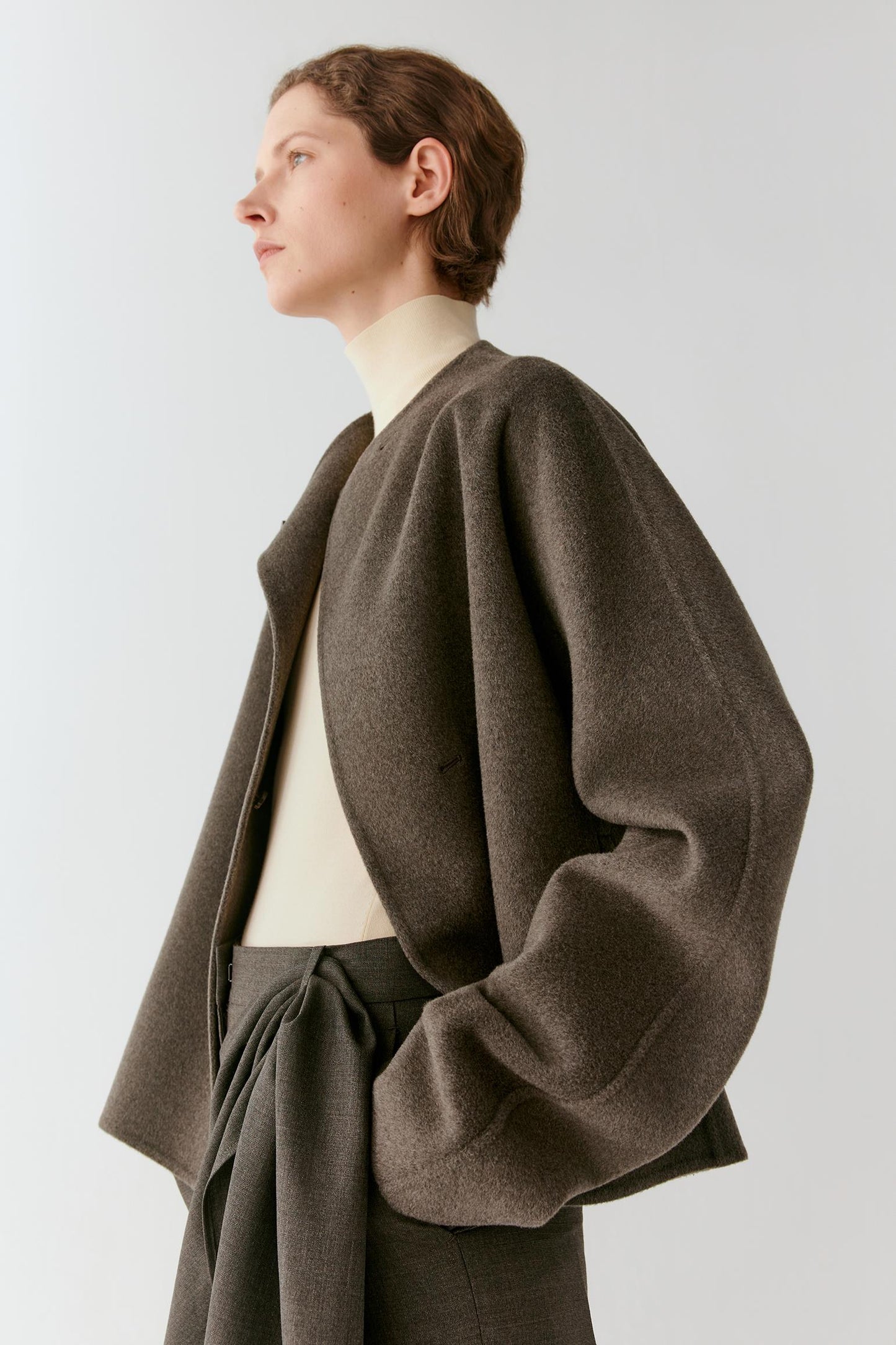 A model in profile wearing a dark brown oversized cropped jacket over a cream turtleneck and dark gray trousers, set against a plain light gray studio background.