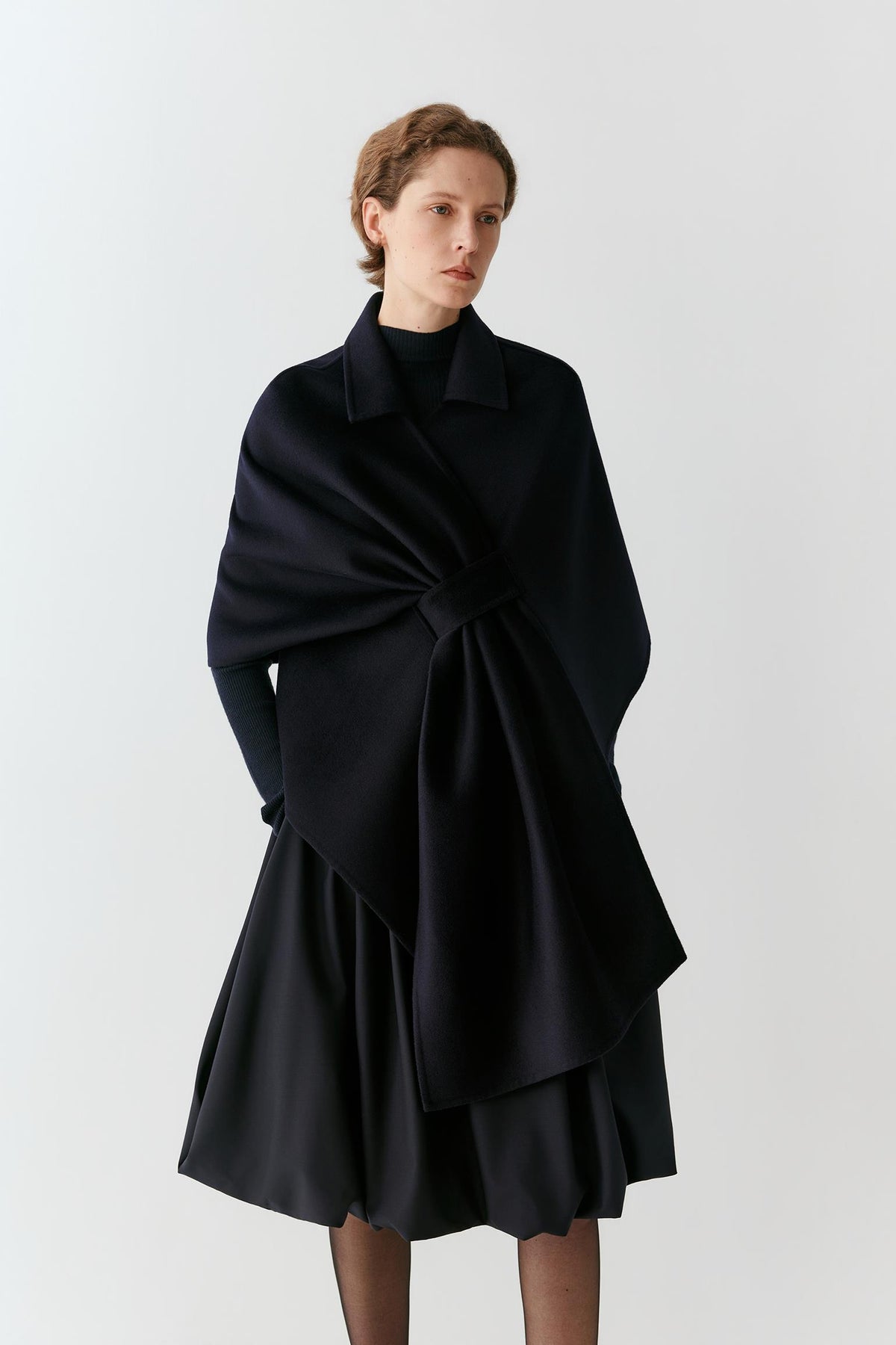 A model wearing a black wrap-style cape with a large bow detail at the waist, paired with a matching black pleated skirt, against a plain light gray studio background.