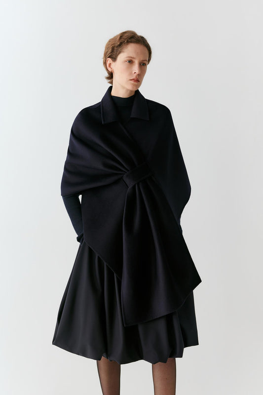 A model wearing a black wrap-style cape with a large bow detail at the waist, paired with a matching black pleated skirt, against a plain light gray studio background.